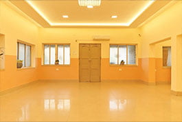 This is a photograph of an extensive, sufficiently bright lobby inside Khelaghar Baganbari, highlighting gleaming floors, light yellow walls with a peach line, and a few windows allowing in regular light. The roof is embellished with brilliant lights and a crystal fixture, and there's a climate control system on the wall.
