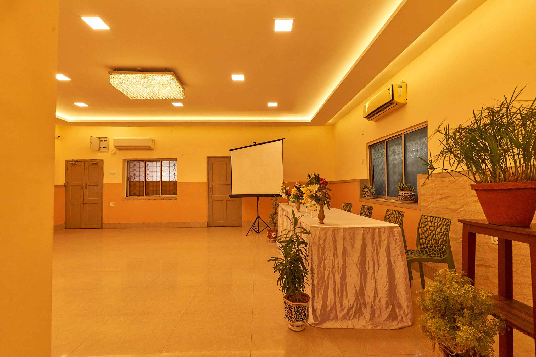 A sufficiently bright, extensive room at Khelaghar Baganbari set up for a gathering or occasion, including a screen for introductions, a table with a flower focal point, agreeable seats, and pruned plants improving the warm feeling.