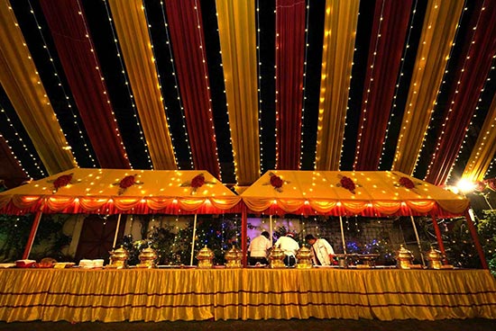 A bubbly open air buffet arrangement around evening time under a covering of yellow and red texture stripes with shimmering lights, serving food in customary metal compartments at Khelaghar Baganbari.