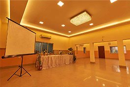 An indoor dinner lobby at Khelaghar Baganbari, enlightened by warm lighting with a projector screen on one side and a long table brightened with plants and blossoms, prepared for an occasion.
