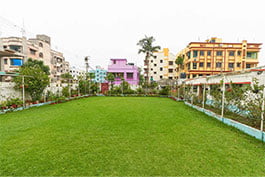An open, green grass lined with pruned plants, catching the quiet and very much kept up with outside space at Khelaghar Baganbari.