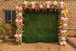 This is a photograph of an embellishing photograph stall with a green support background, outlined by a brilliant game plan of blossoms and inflatables, set up for a festival at Khelaghar Baganbari.