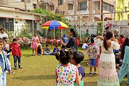 Children are playing on a grassy lawn under a bright umbrella at Khelaghar Baganbari, with adults supervising in the background.