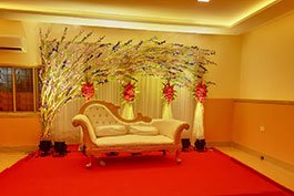 A luxurious wedding couch set under an enriching curve with purple blossoms and red accents, situated on a phase with red covering, exhibiting Khelaghar Baganbari's rich occasion styling.
